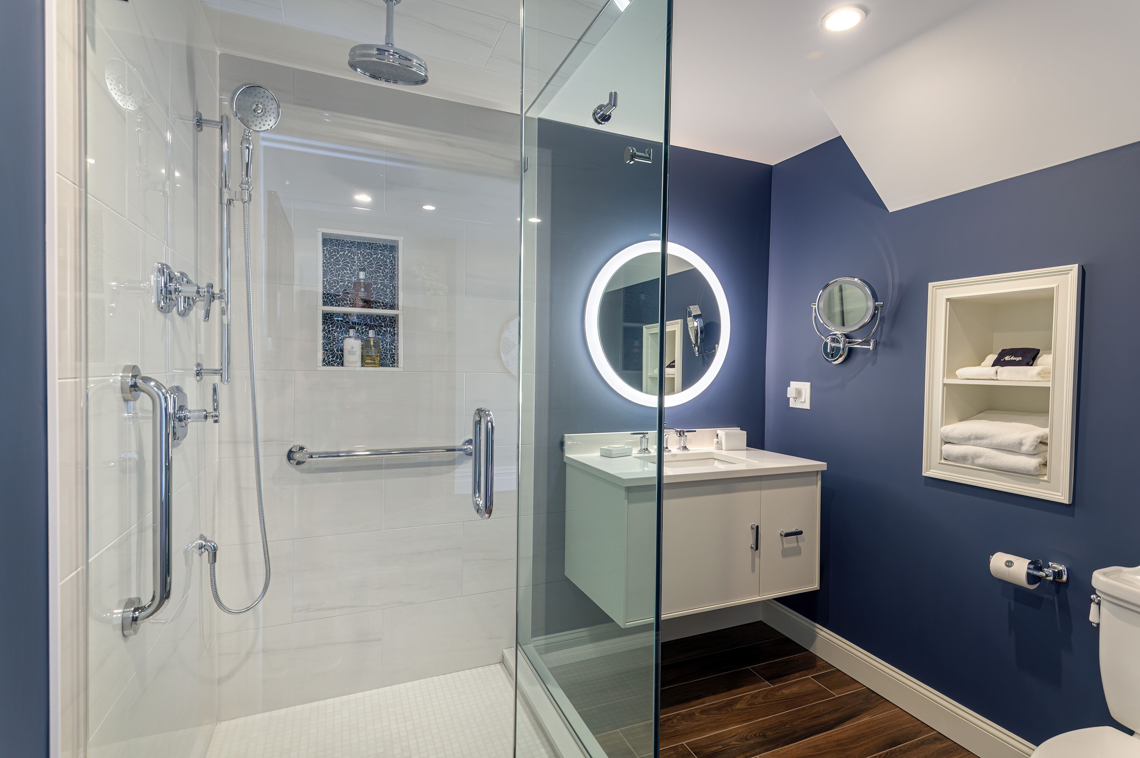 A modern bathroom with a glass-enclosed shower, a round mirror with a light, a white sink, and a white toilet.