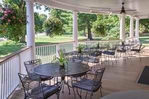A covered outdoor patio with round tables and chairs, surrounded by white columns, and overlooking a well-maintained lawn and trees.