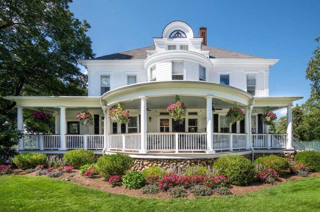A large, white, two-story house with a wrap-around porch and hanging flower baskets.