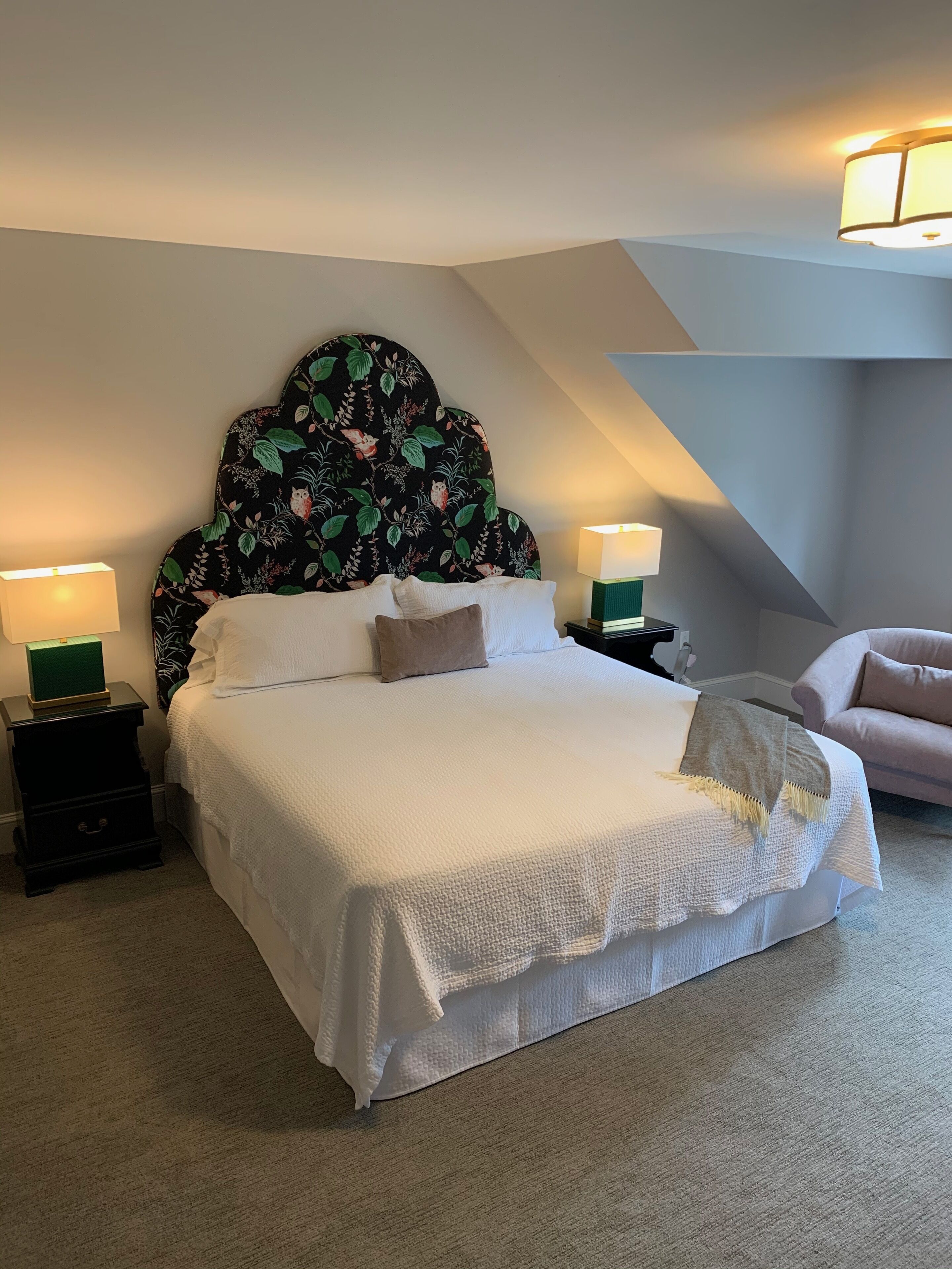 A neatly made bed with a floral headboard, two bedside tables with lamps, and a chair in a modern room with a sloped ceiling.