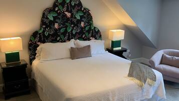 A neatly made bed with a floral headboard, two bedside tables with lamps, and a chair in a modern room with a sloped ceiling.