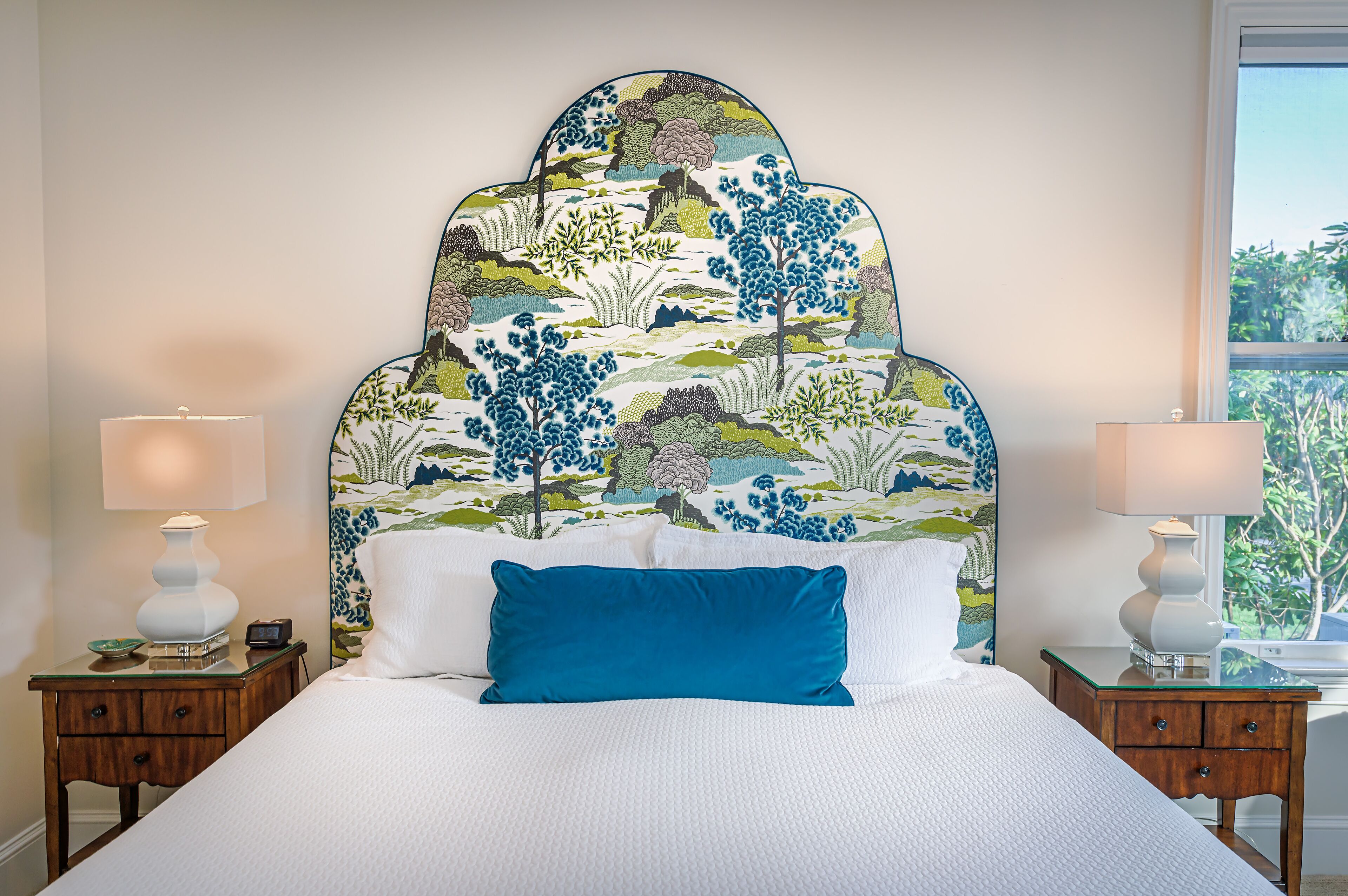 Una cama con una colcha blanca y almohada azul, flanqueada por dos mesitas de noche con lámparas, en una habitación con un cabecero estampado y vistas al exterior a través de una ventana.