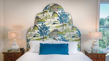 Una cama con una colcha blanca y almohada azul, flanqueada por dos mesitas de noche con lámparas, en una habitación con un cabecero estampado y vistas al exterior a través de una ventana.