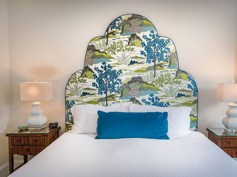 Una cama con una colcha blanca y almohada azul, flanqueada por dos mesitas de noche con lámparas, en una habitación con un cabecero estampado y vistas al exterior a través de una ventana.
