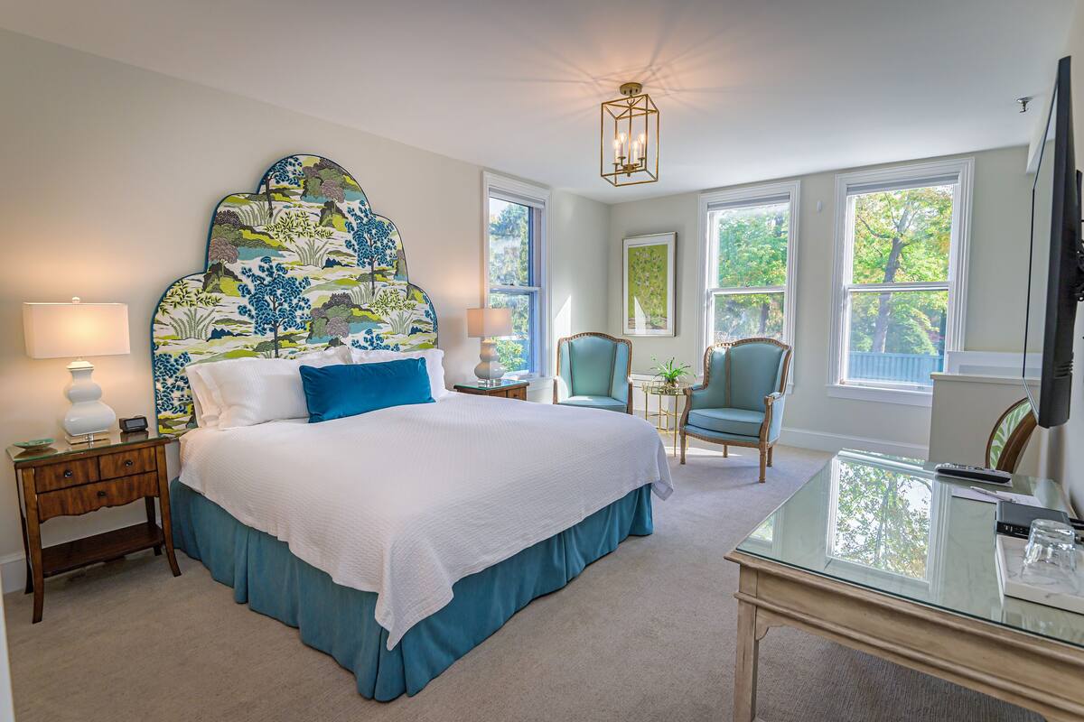 A bedroom with a large bed, a desk, and a chair. There are windows and a ceiling light fixture.