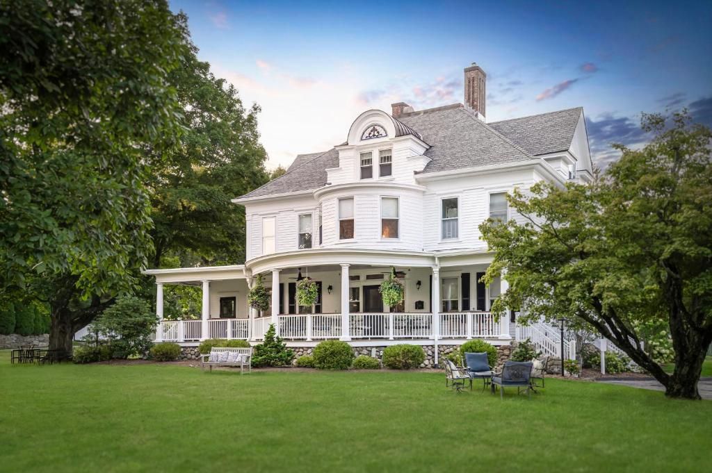 Une grande maison blanche à deux étages, dotée d’une véranda qui fait le tour de la maison, et entourée d’une pelouse bien entretenue et d’arbres.
