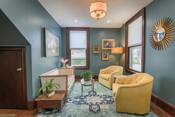A room with a blue wall, a wooden door, a white sofa, a yellow armchair, a small table with a plant, and a patterned rug.