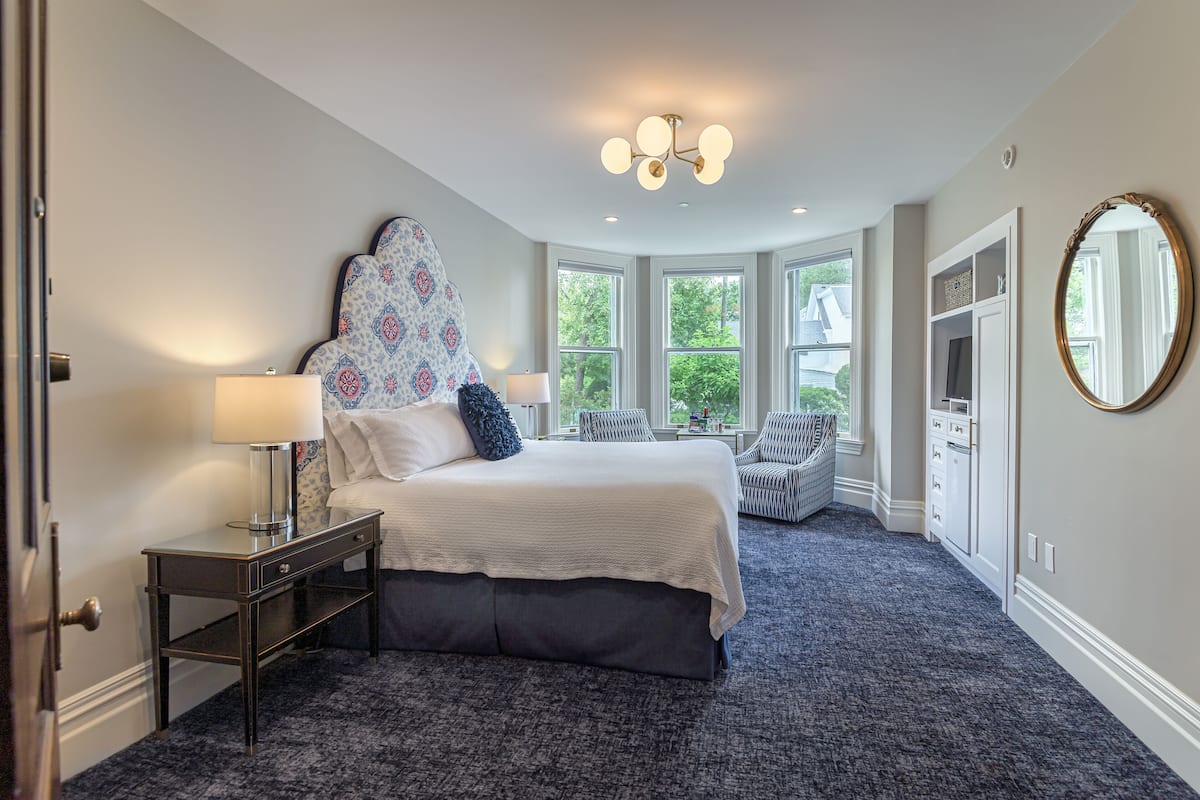 A bedroom with a large bed, two bedside tables, a dresser, and a mirror. There are windows providing natural light and a view of greenery outside.