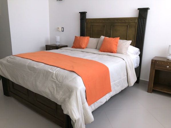 Una cama con cabecera de madera, ropa de cama blanca y naranja, y una mesita de noche con una lámpara.