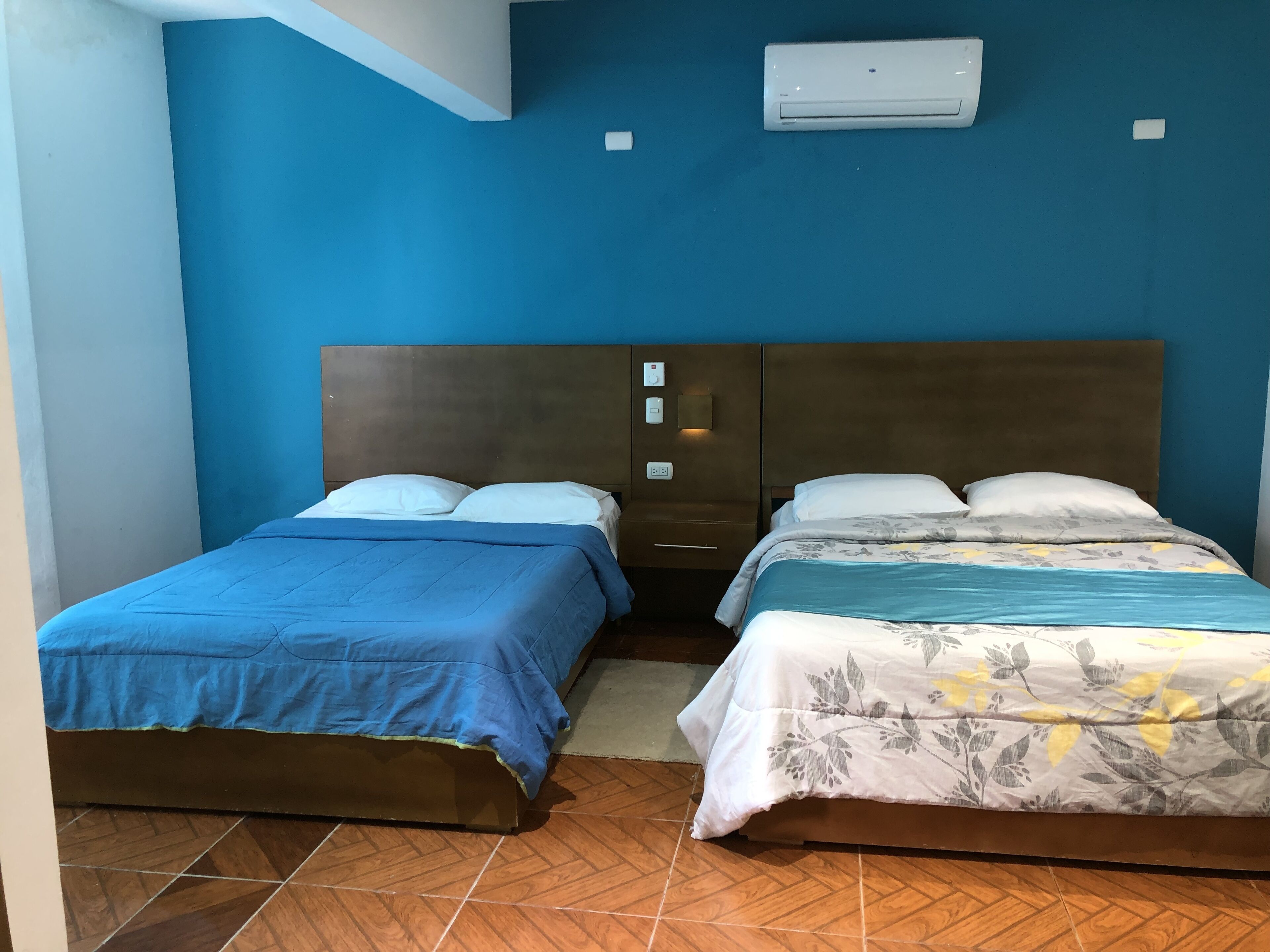 A room with two beds, each with a different style of bedding, a wooden headboard, and a wall-mounted air conditioning unit.