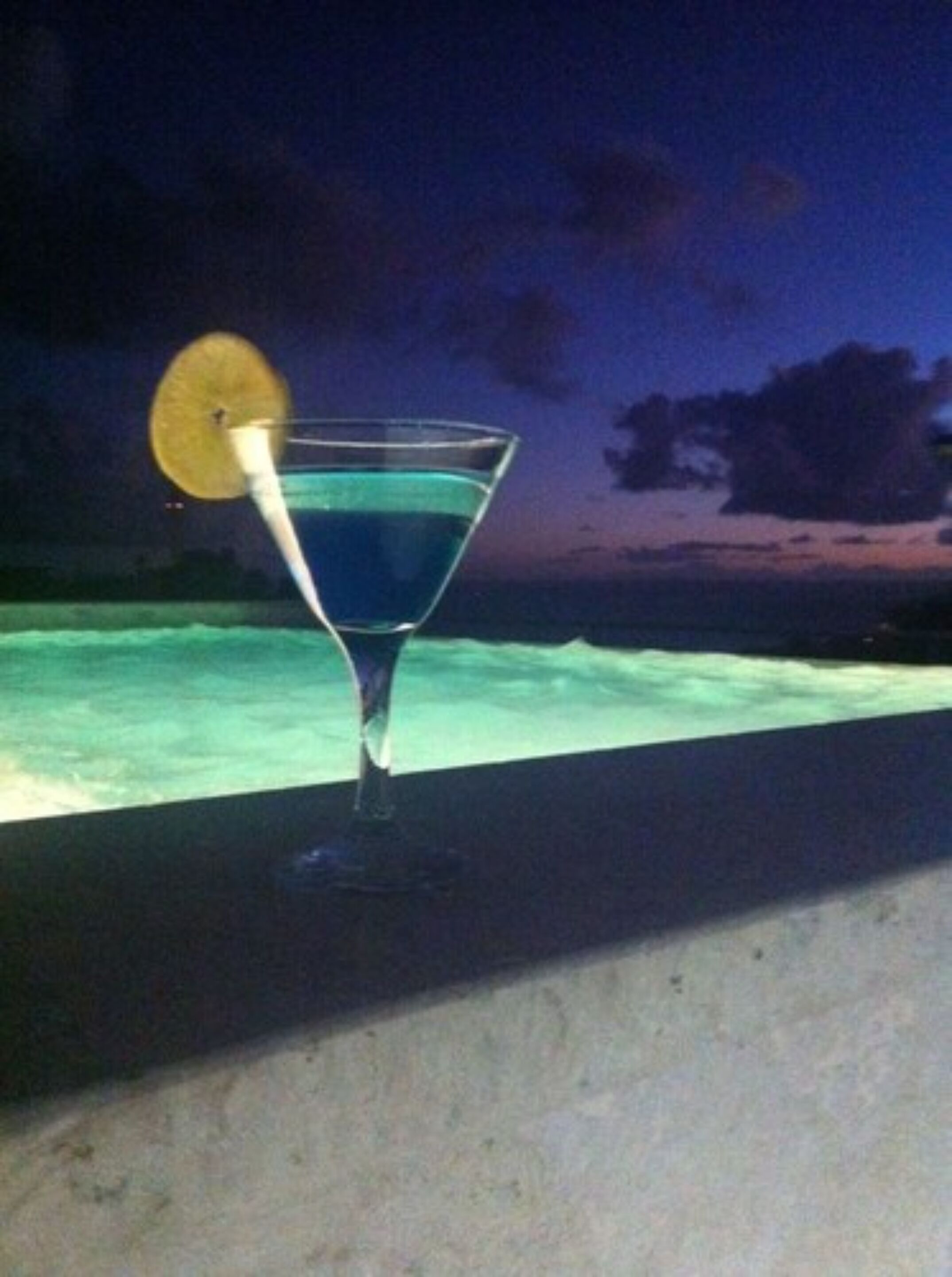 A martini glass with a lemon slice on a balcony overlooking a body of water at night.