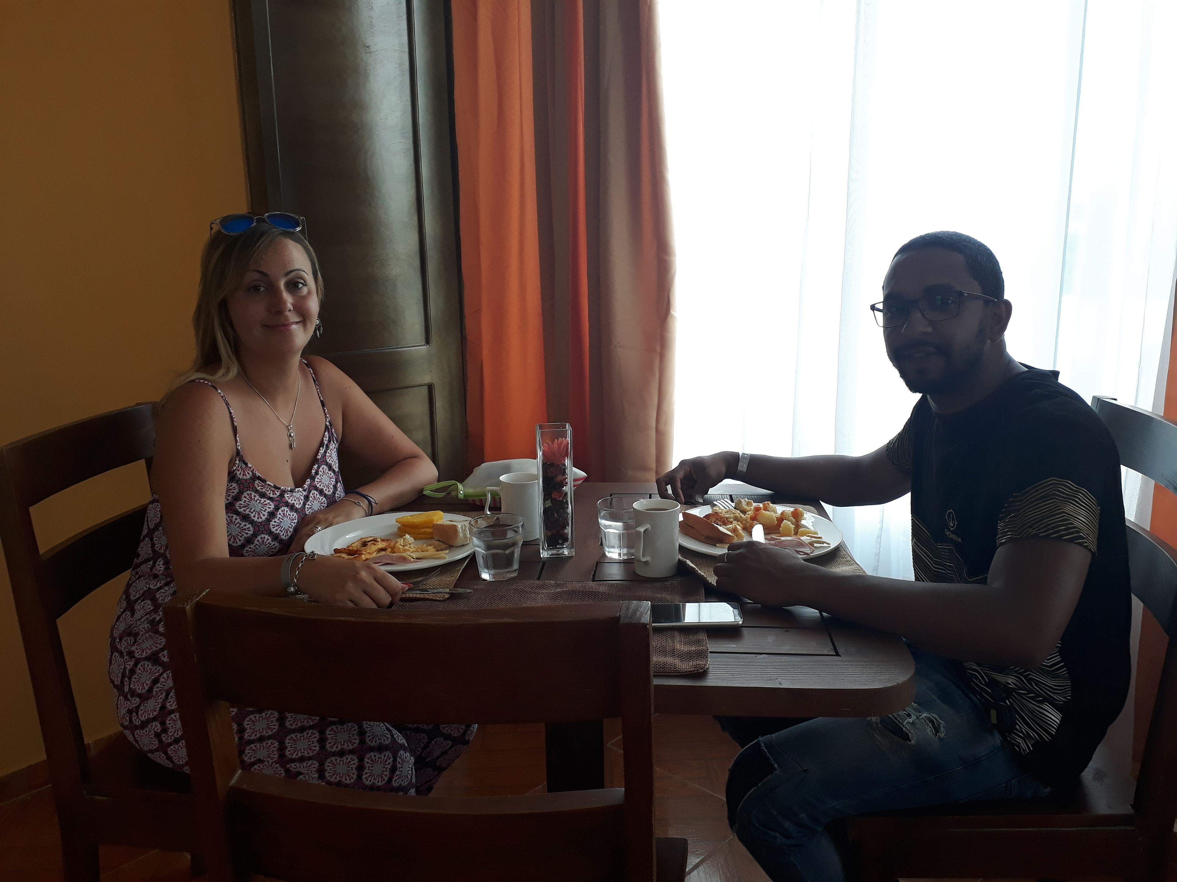 Dos personas sentadas en una mesa de madera con platos de comida, vasos y un teléfono inteligente.
