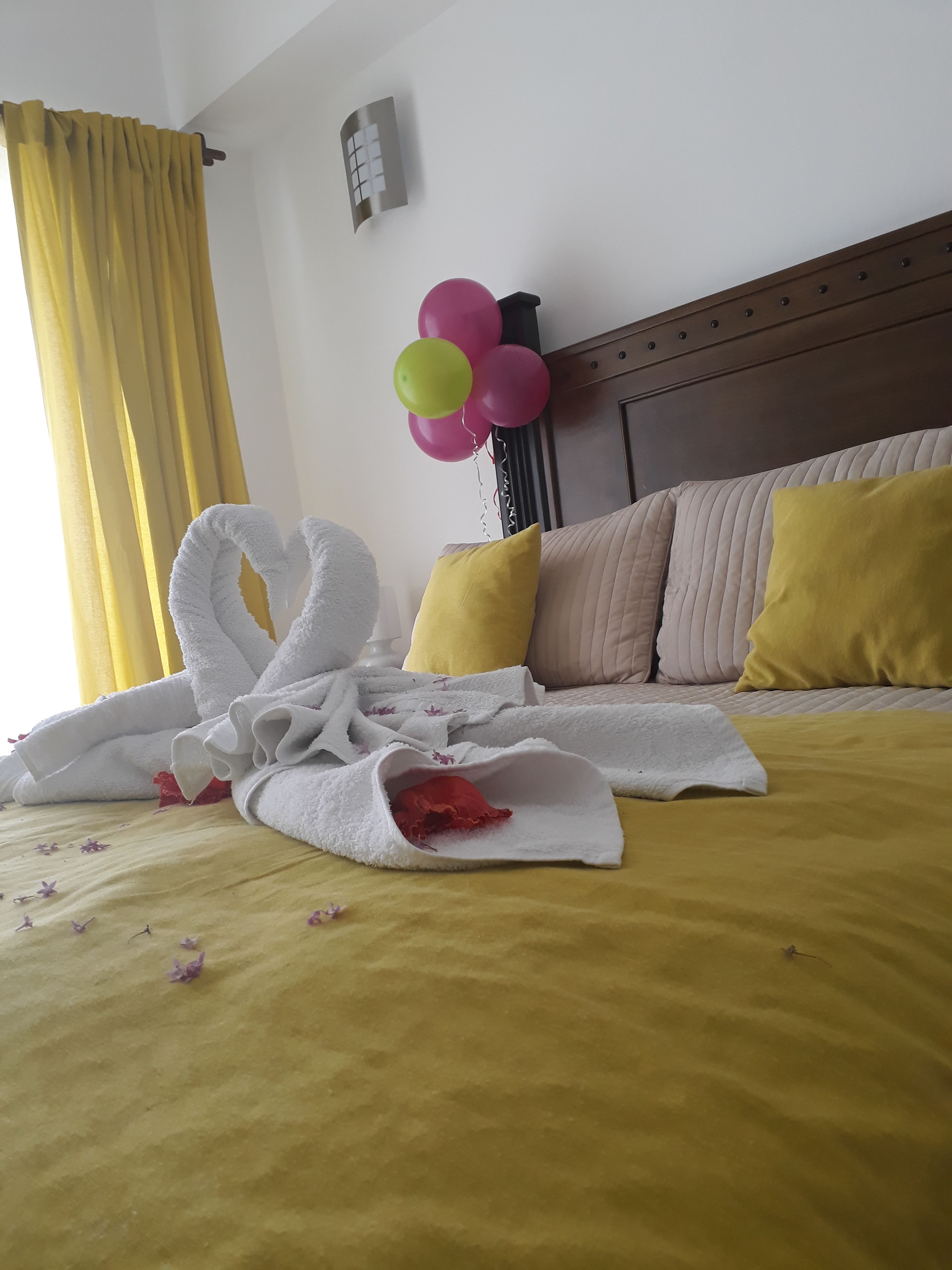A neatly made bed with a yellow bedspread, a white towel arranged in a heart shape, and a bouquet of balloons in pink, yellow, and green.