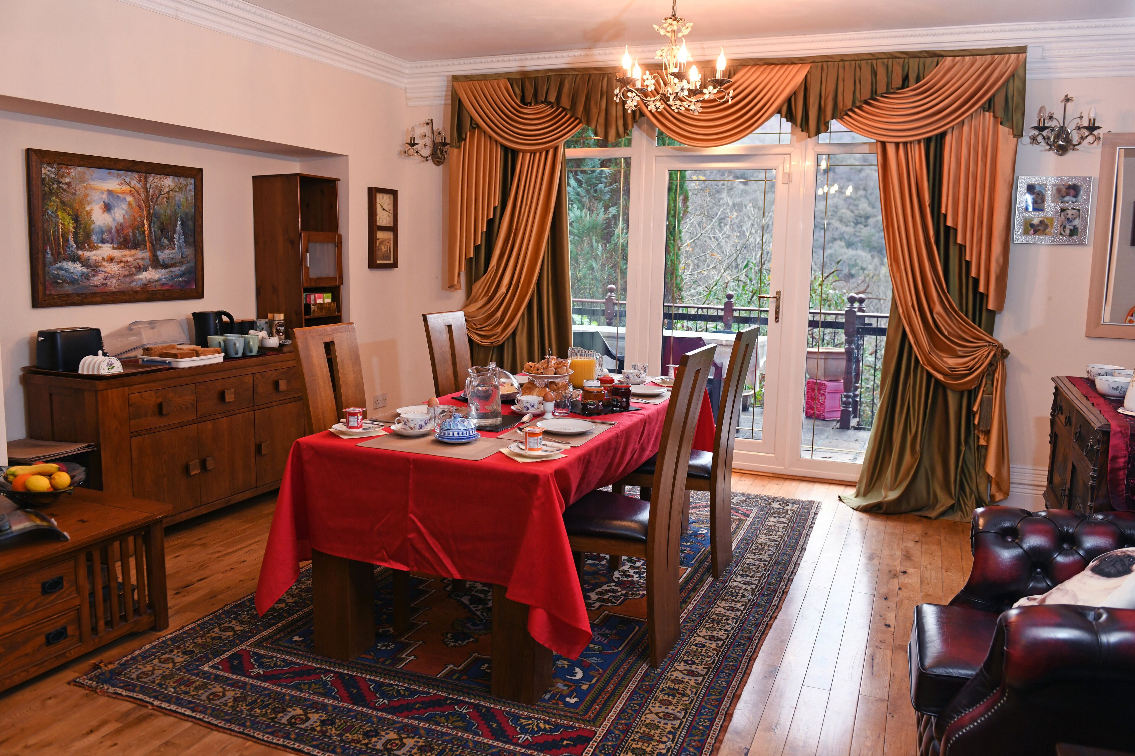 A dining room with a red tablecloth, wooden chairs, and a chandelier. There is a painting on the wall and a window with curtains.