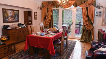 Comedor con mantel rojo, sillas de madera y candelabro. Hay un cuadro en la pared y una ventana con cortinas.
