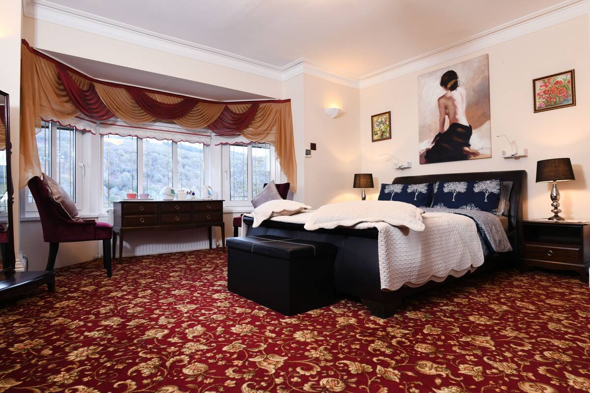 Une chambre avec un grand lit, un bureau et une chaise. Il y a une fenêtre avec des rideaux, un tableau au mur et une lampe sur la table de chevet.