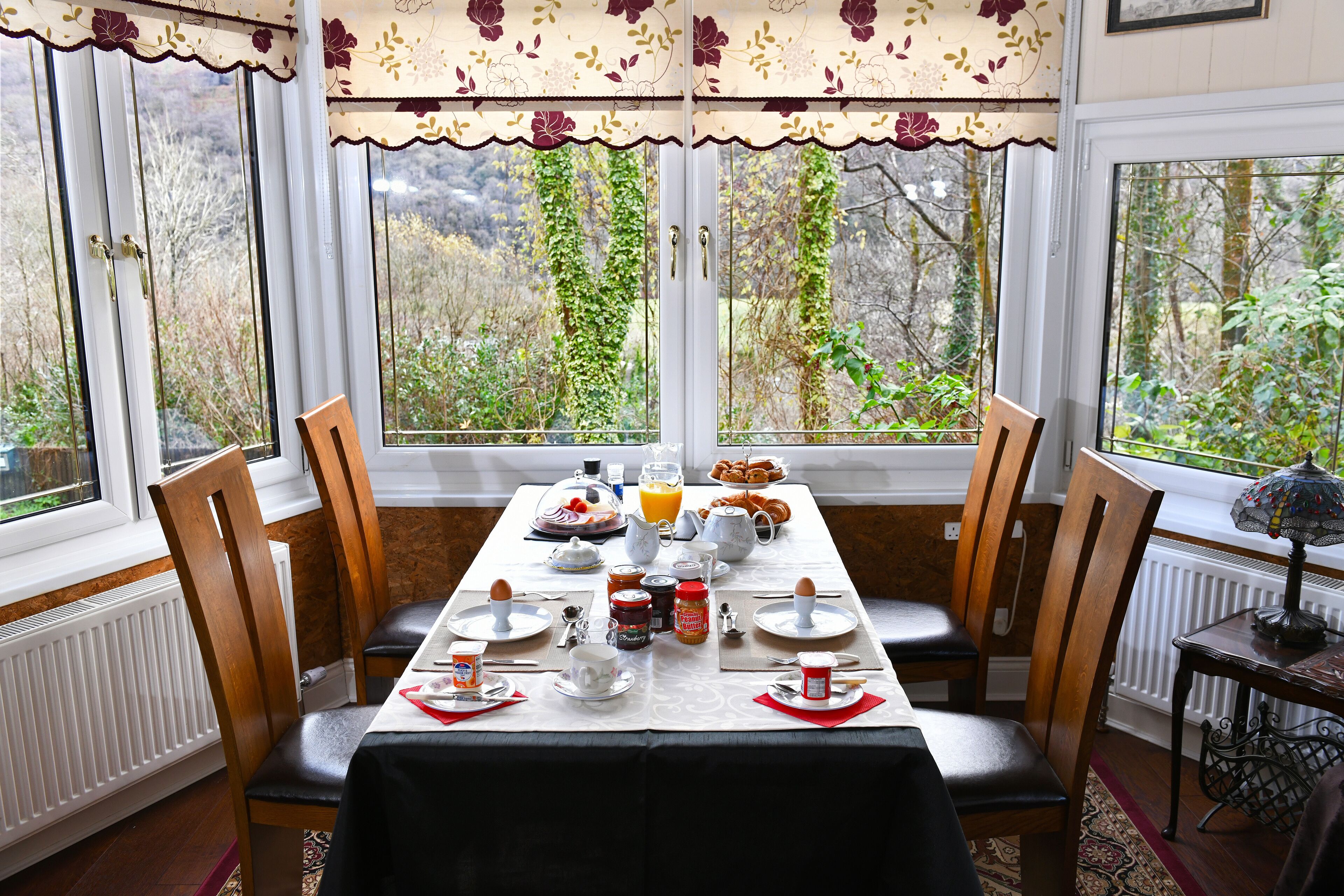 Una mesa de desayuno dispuesta con platos, tazas y diversos alimentos para el desayuno, rodeada de sillas, con vistas a la vegetación a través de amplias ventanas.