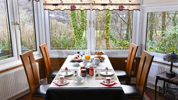 A breakfast table set with plates, cups, and various breakfast items, surrounded by chairs, with a view of greenery through large windows.