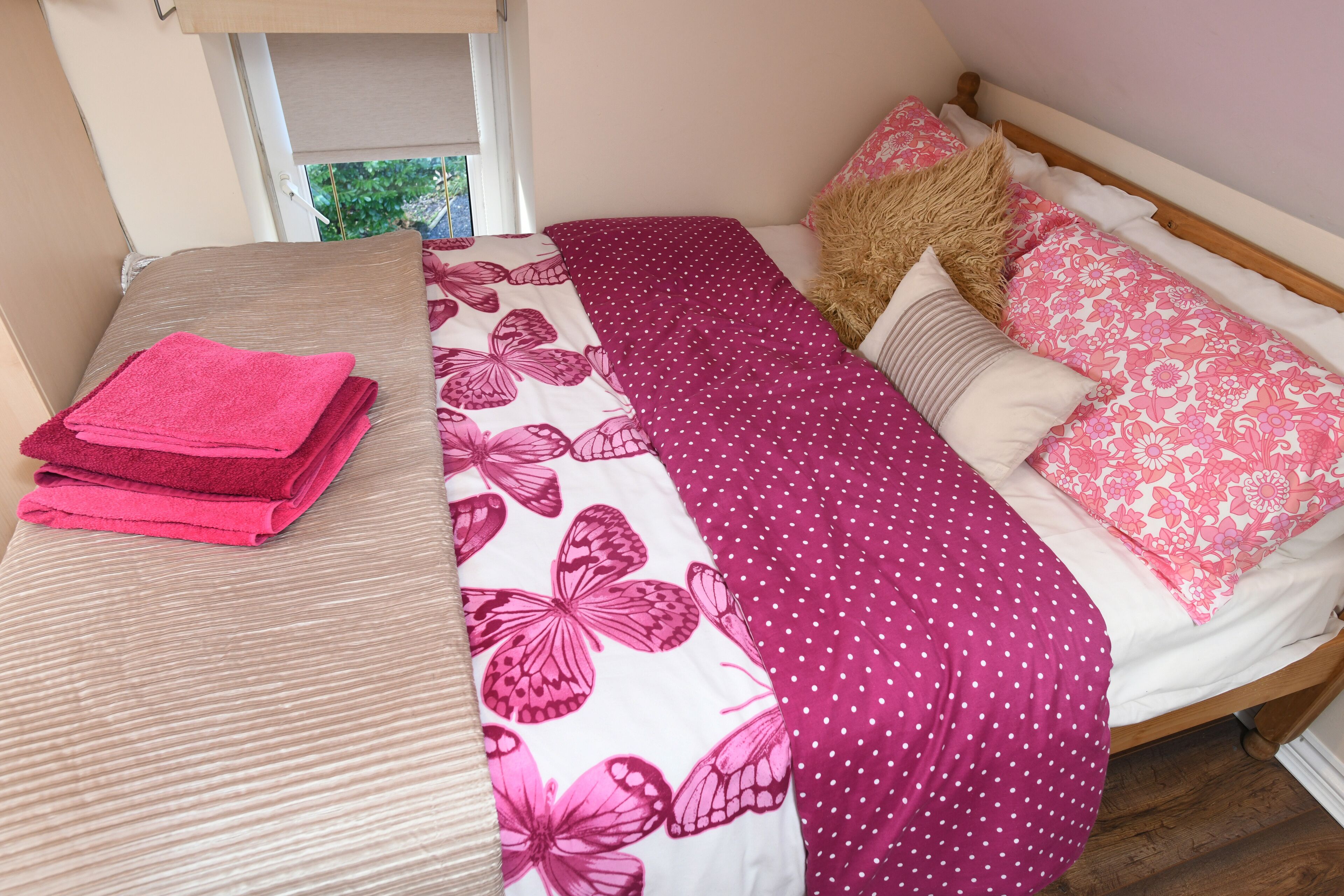 Una cama bien hecha con ropa de cama floral en rosa y blanco, un cabecero de madera y una ventana con vistas a follaje.