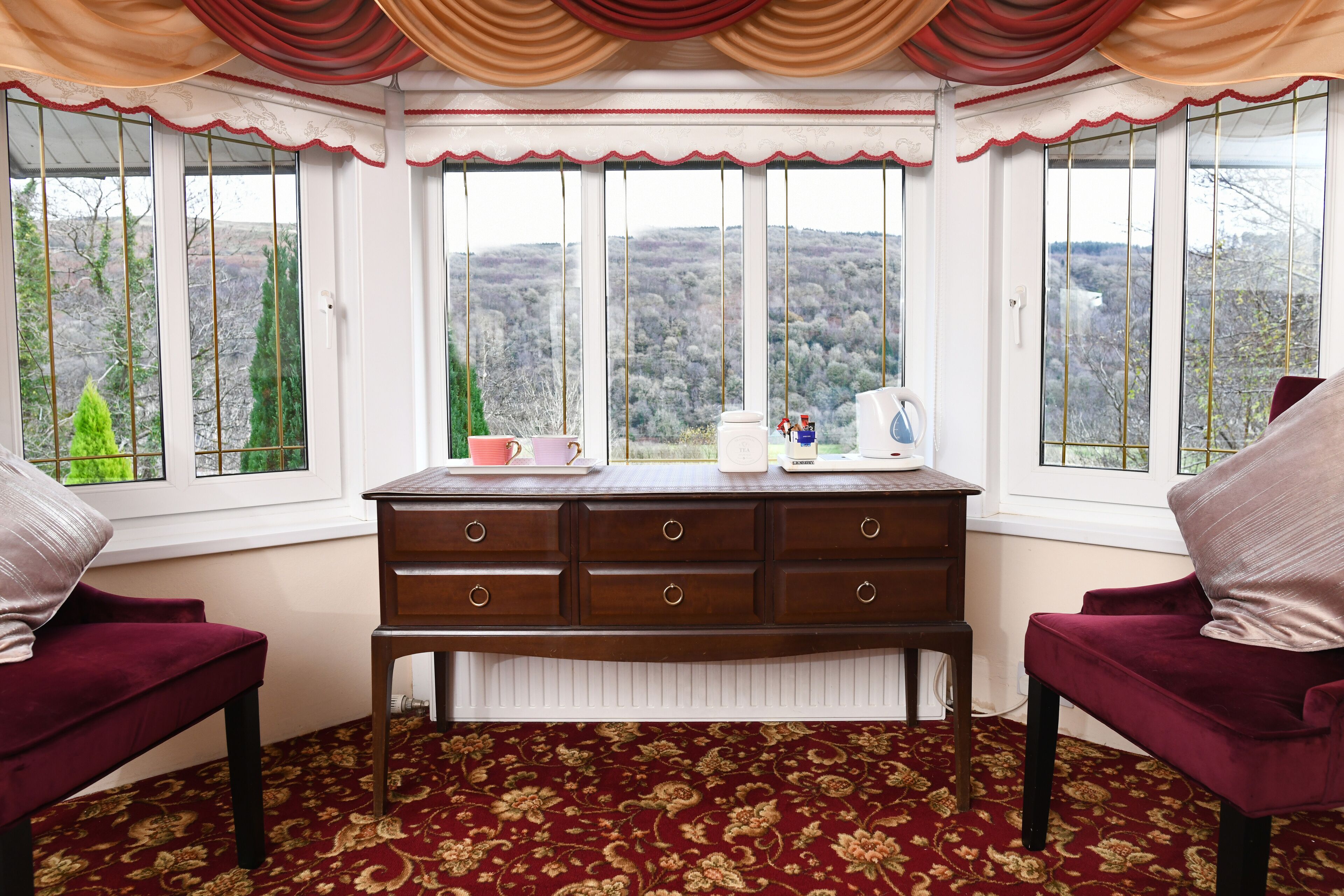 A room with a large bay window, a wooden dresser, two red armchairs, and a patterned carpet.