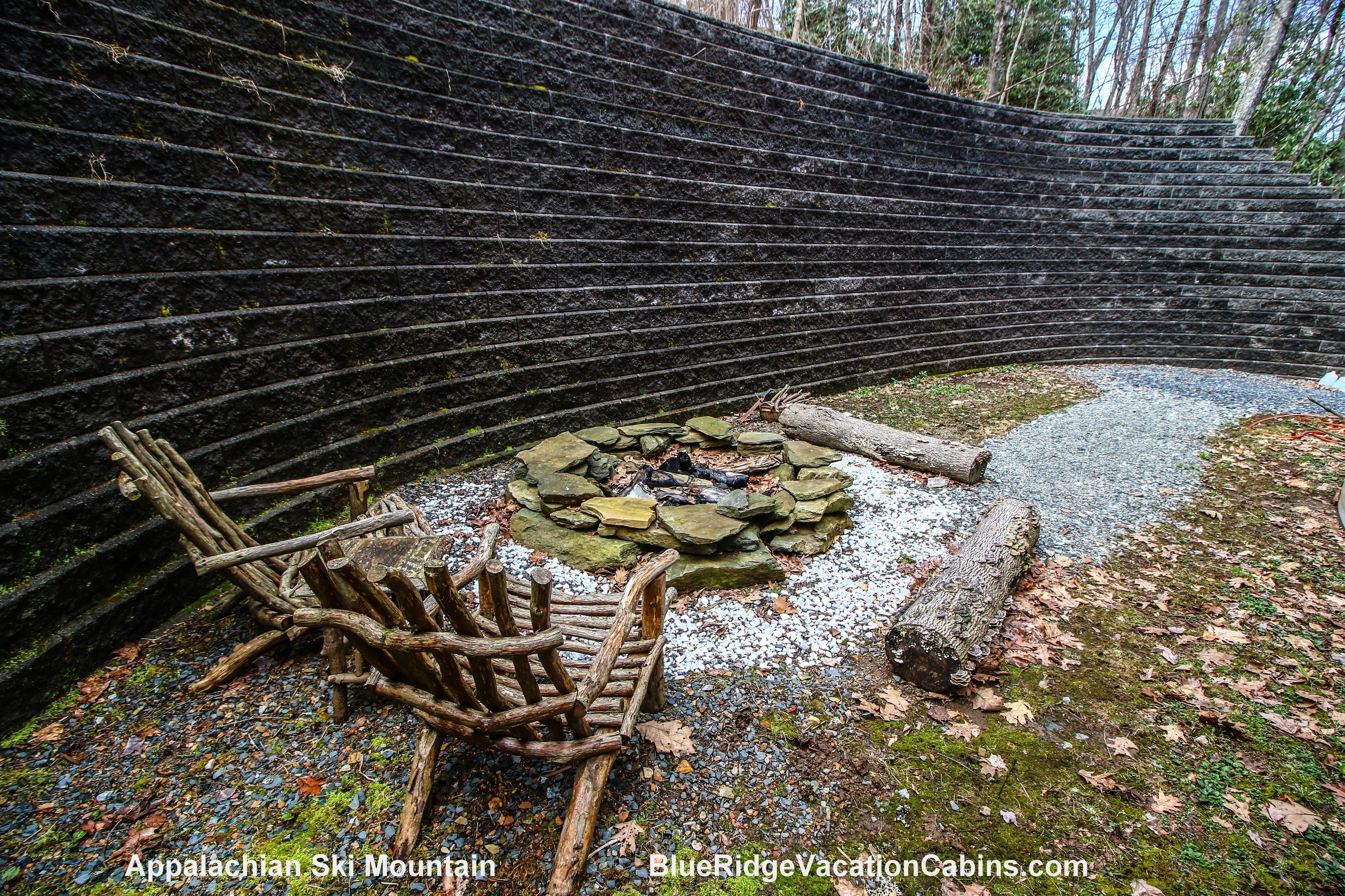 A rustic outdoor seating area with a stone fire pit, surrounded by a stone wall and natural forest backdrop.