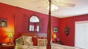 Een hemelbed met plafondventilator, een houten vloer en een rode muur met een spiegel en ingelijste foto's.