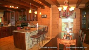 A cozy lodge kitchen with wooden beams, a stone island, bar stools, and a chandelier. - Lodge at Riversound Apartment (Banner Elk)