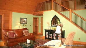 A cozy living room with a wooden ceiling, a brown leather sofa, a glass coffee table, and a bookshelf. - Lodge at Riversound Apartment (Banner Elk)