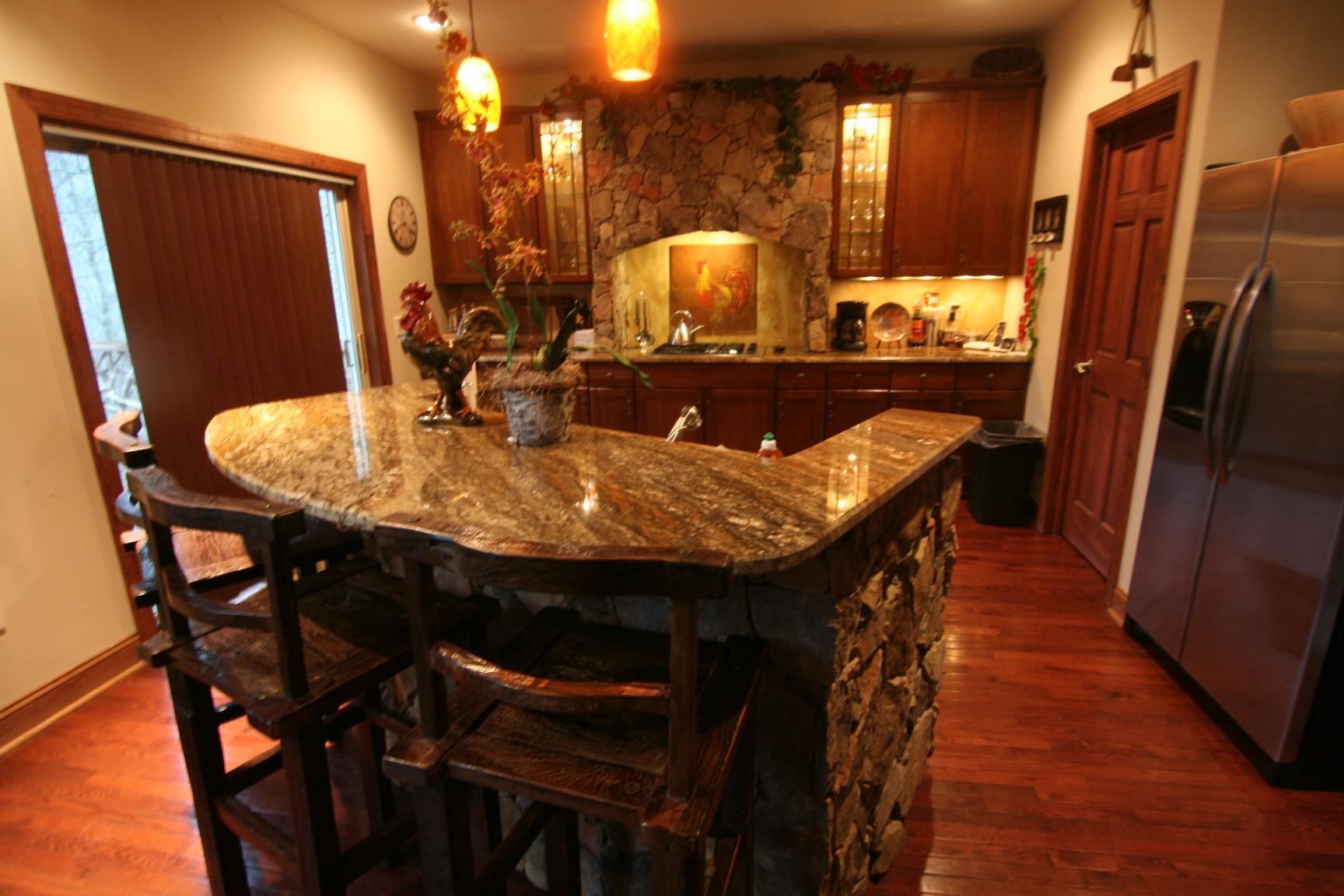 A kitchen with a stone accent wall, wooden cabinets, a granite island with bar stools, and a stainless steel refrigerator.