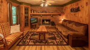 A cozy living room with a fireplace, wooden walls, a ceiling fan, and a television.