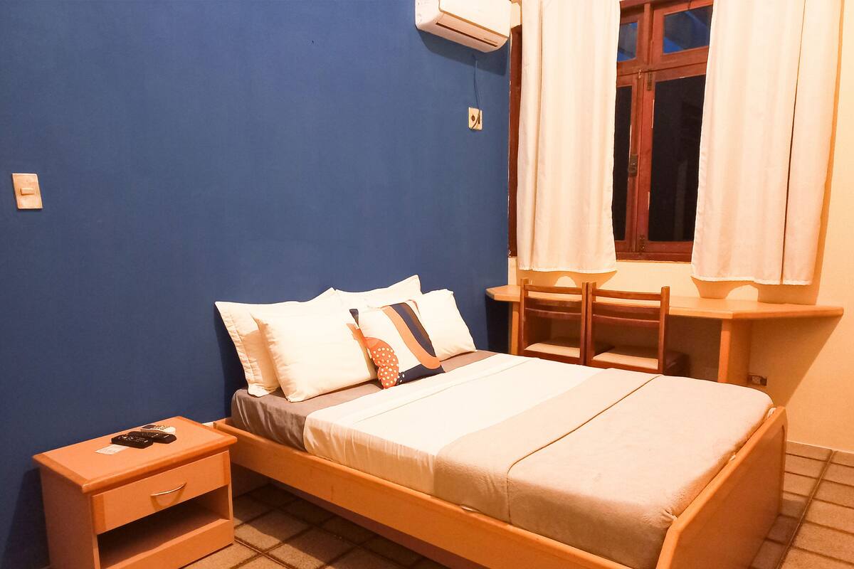 A single bed with a wooden frame, a nightstand with a drawer, a small table, and a window with curtains.