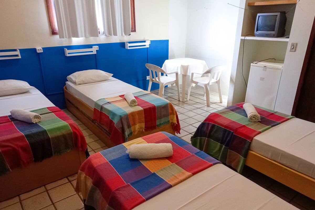 A hotel room with three beds, each with a colorful blanket and a rolled towel. There is a small table with a white tablecloth and two white chairs. A television and a refrigerator are visible in the corner.