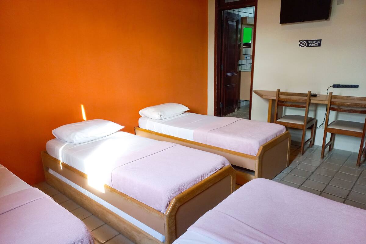 A room with three wooden beds with white bedding, an orange wall, a wooden desk with chairs, and a television on the wall.