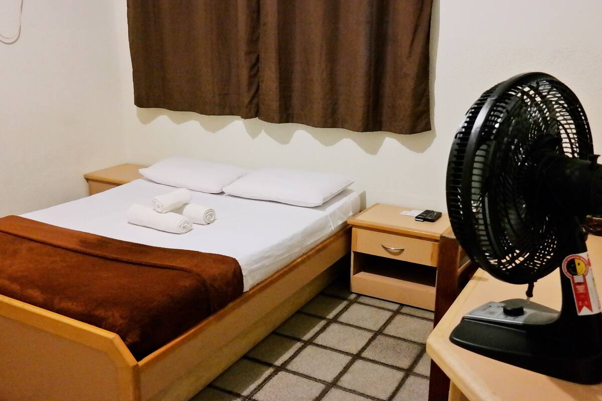 A single bed with a brown blanket, white linens, and two rolled towels. A bedside table with a remote control and a fan is next to the bed.
