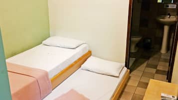 A small, single-bed room with a wooden frame, white bedding, and a pink towel on the left side. A sink and toilet are visible in the adjacent bathroom.