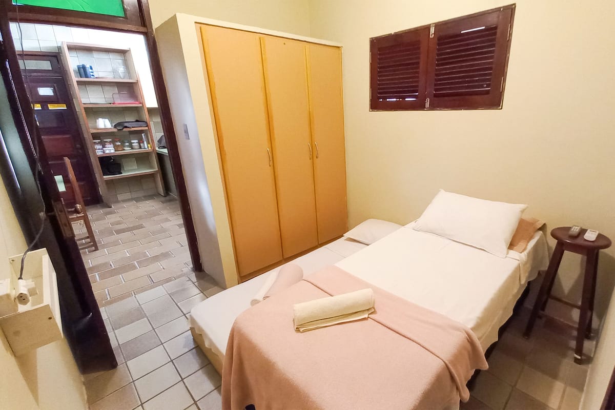 A bedroom with a bed, a wardrobe, a small stool, and a window with blinds.