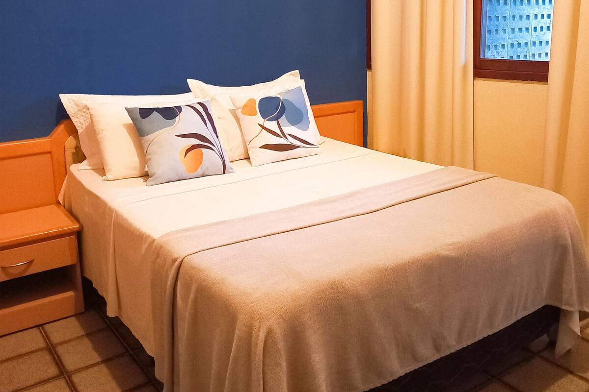 A neatly made bed with a white bedspread and decorative pillows, a wooden nightstand with a drawer, and a window with curtains.