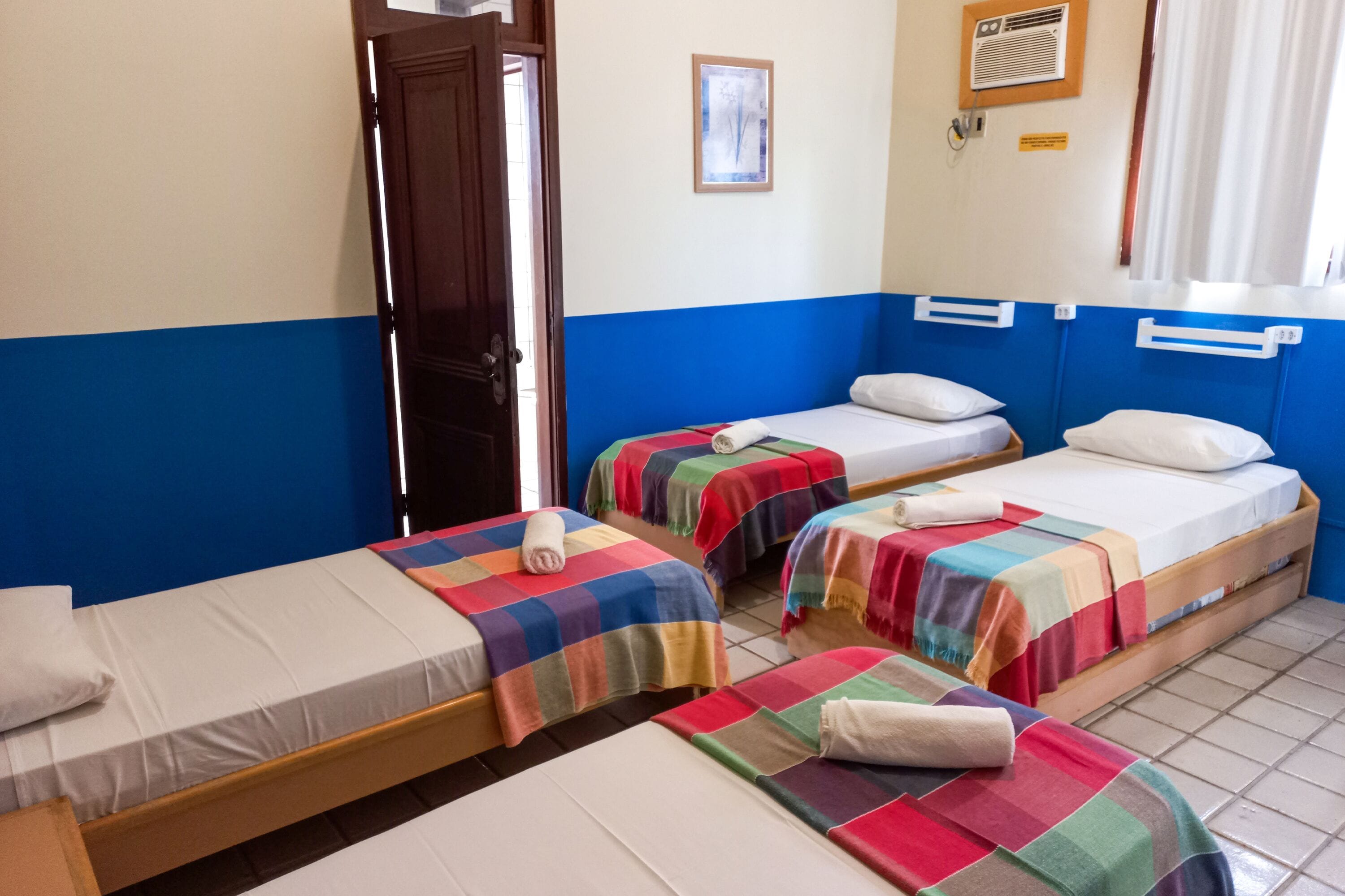 Una habitación con tres camas individuales, cada una con una manta colorida y un almohadón blanco. Se observa una puerta de madera a la izquierda, un cuadro enmarcado en la pared y una ventana con cortinas.