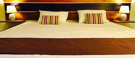 Cama bem arrumada com roupa de cama branca e almofadas listradas, cabeceira de madeira e duas luminárias de mesa.