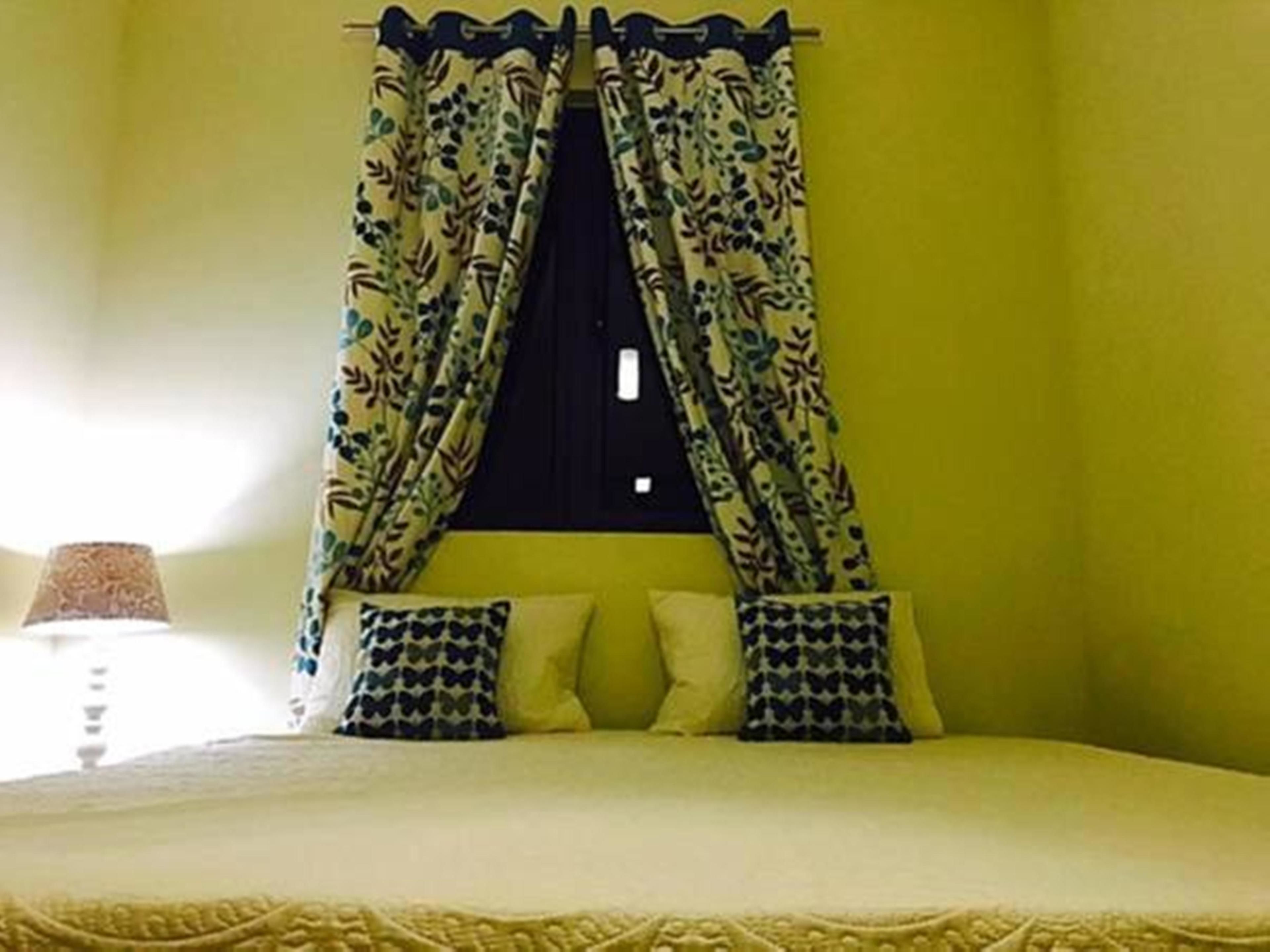 Een kamer met een bed, een raam met bloemen gordijnen en een lampje op een nachtkastje.