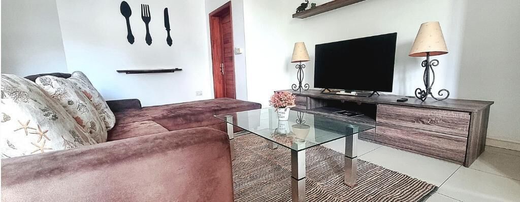 A living room with a glass coffee table, a sofa, a TV on a wooden stand, and two lamps.