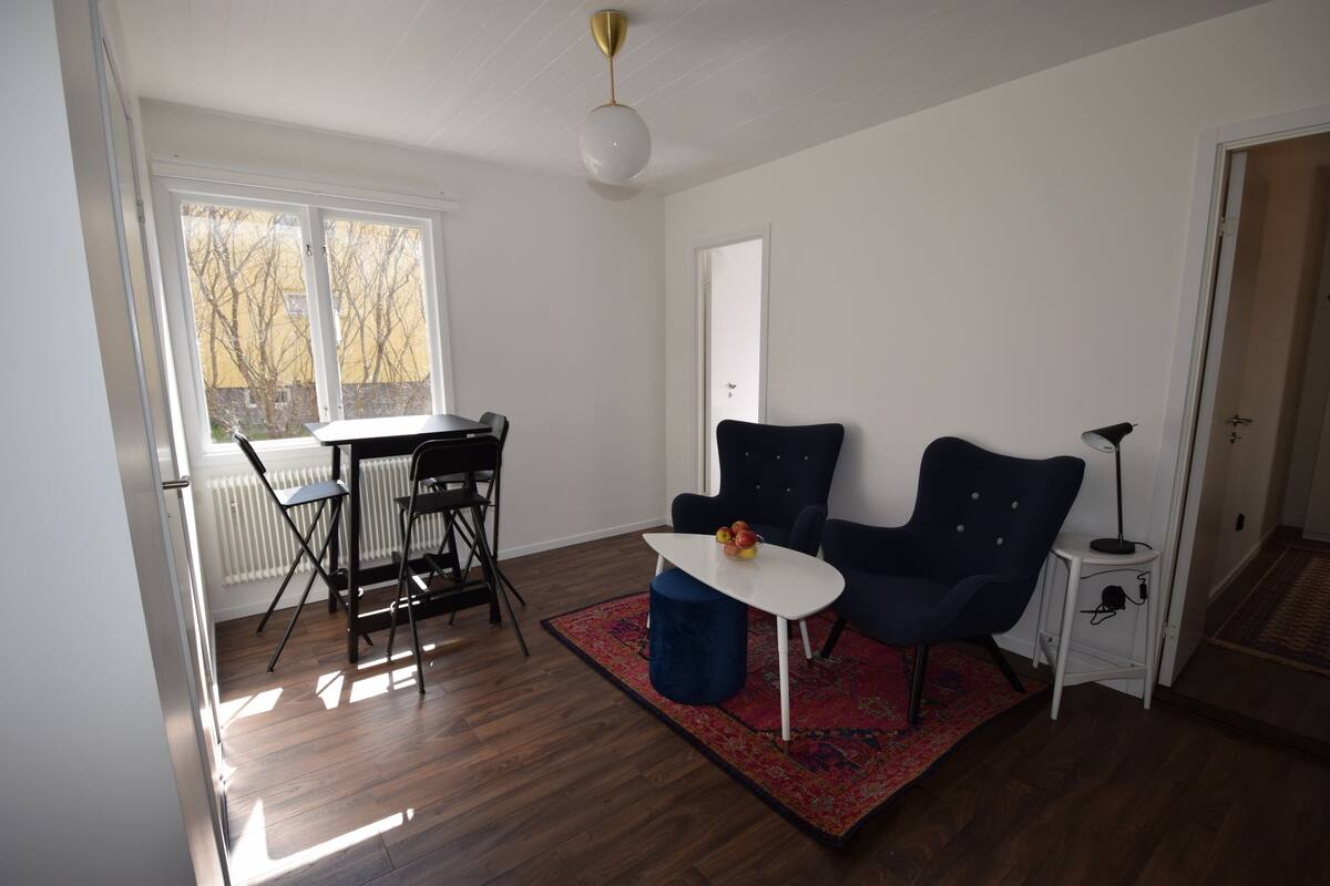 A room with a dining table, two chairs, a small round table with a bowl, a blue ottoman, a floor lamp, and a radiator.