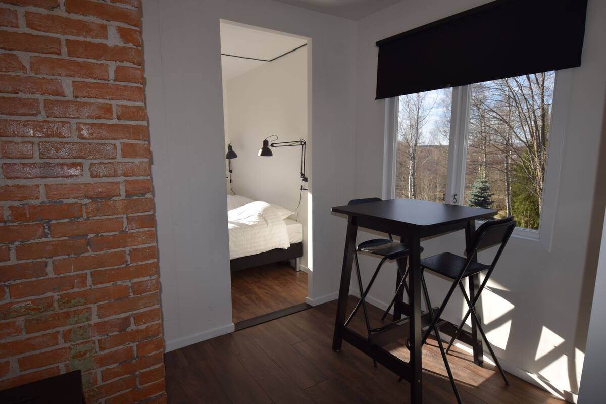 A room with a brick wall, a bed with a white headboard, a desk with a chair, and a window with a view of trees.