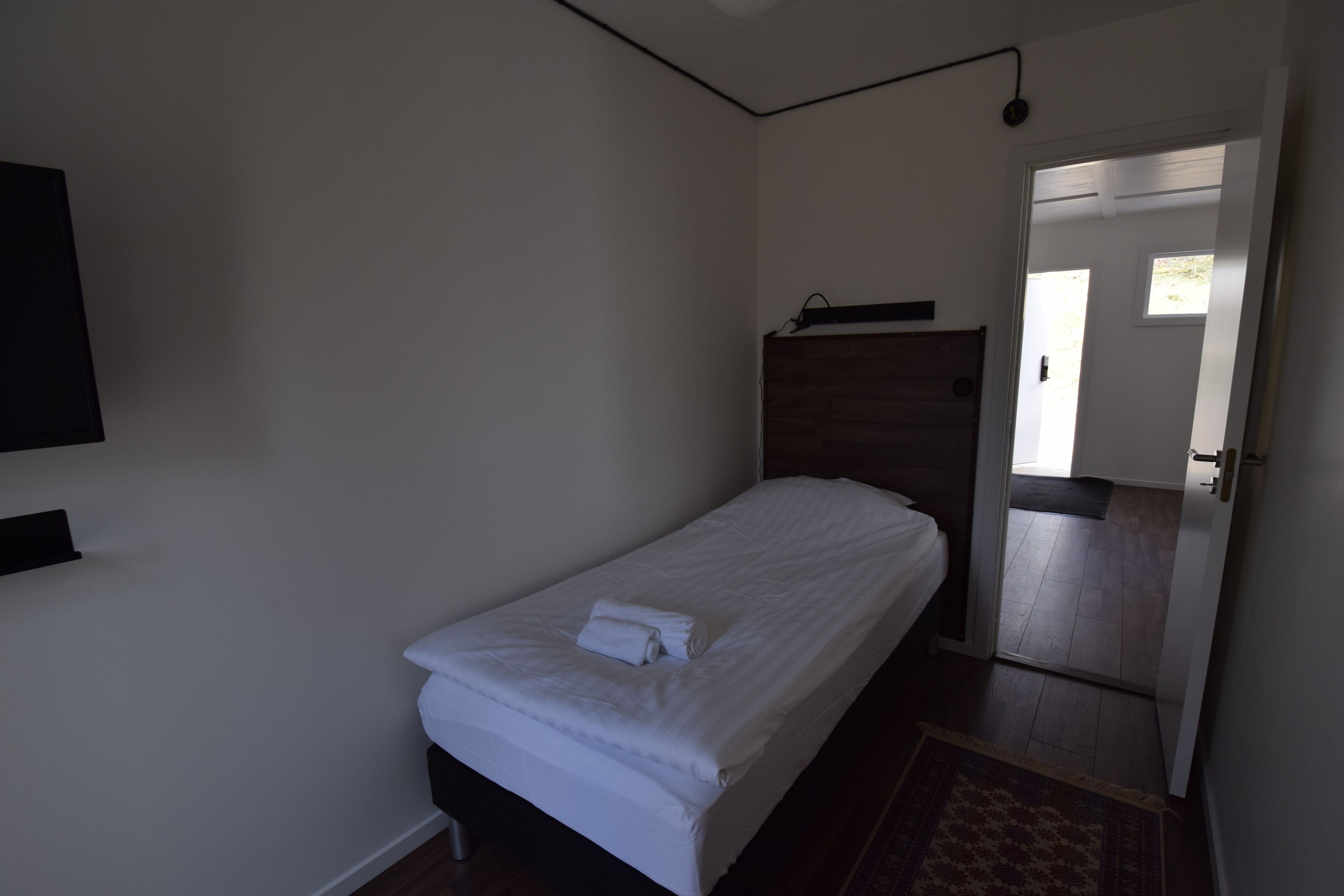 Una cama individual con ropa de cama blanca y una toalla enrollada, un cabecero de madera, un televisor de pantalla plana montado en la pared y una puerta que da a un pasillo.