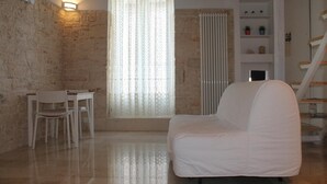 A room with a stone wall, a white chair, a white sofa, a window with a sheer curtain, and a staircase. - B&B Piazza Garibaldi (Santeramo in Colle)