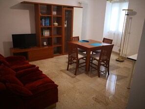 A living room with a red sofa, a wooden dining table with four chairs, a television on a wooden stand, a bookshelf, and a floor lamp. - B&B Piazza Garibaldi (Santeramo in Colle)