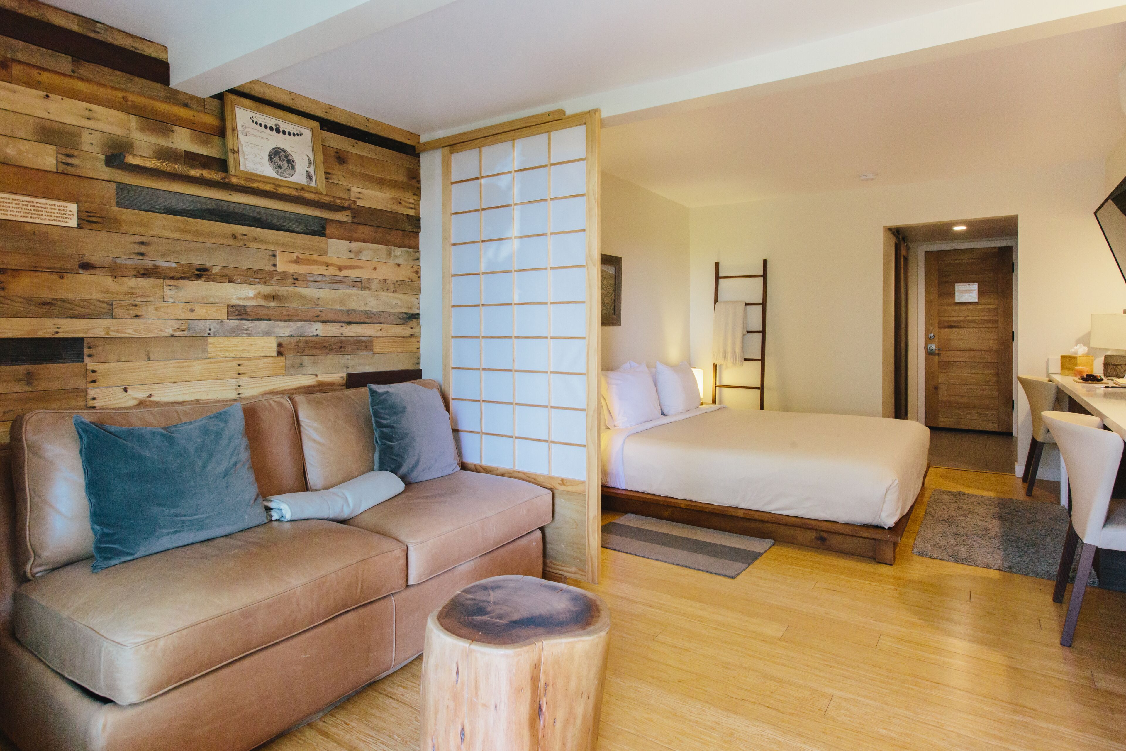 A modern hotel room with a wooden wall, a sofa, a bed, a desk, and a chair.
