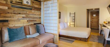 A modern hotel room with a wooden wall, a sofa, a bed, a desk, and a chair.