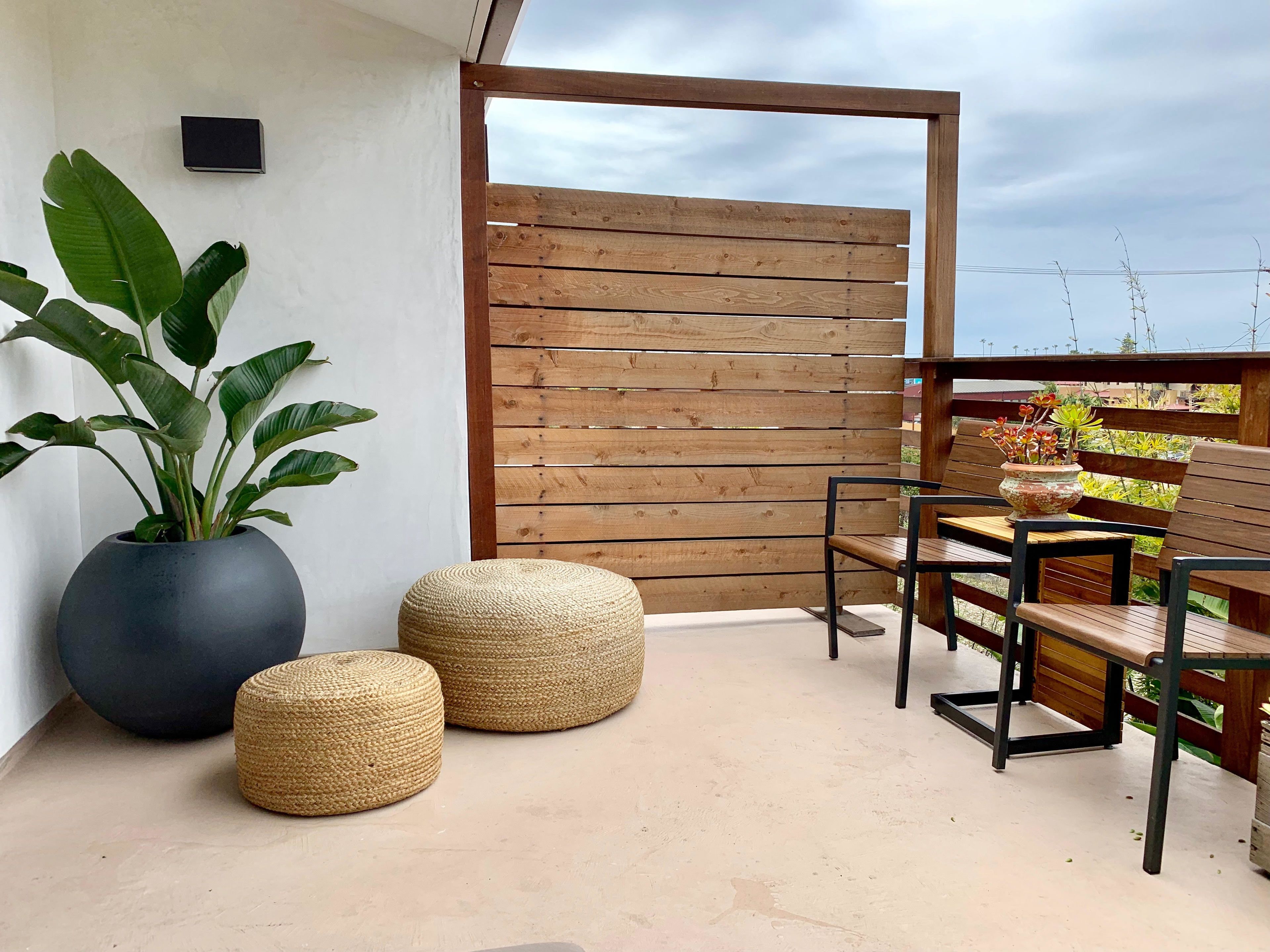 A balcony with a wooden fence, two woven poufs, a large potted plant, and a table with chairs.