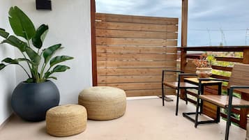 A balcony with a wooden fence, two woven poufs, a large potted plant, and a table with chairs.